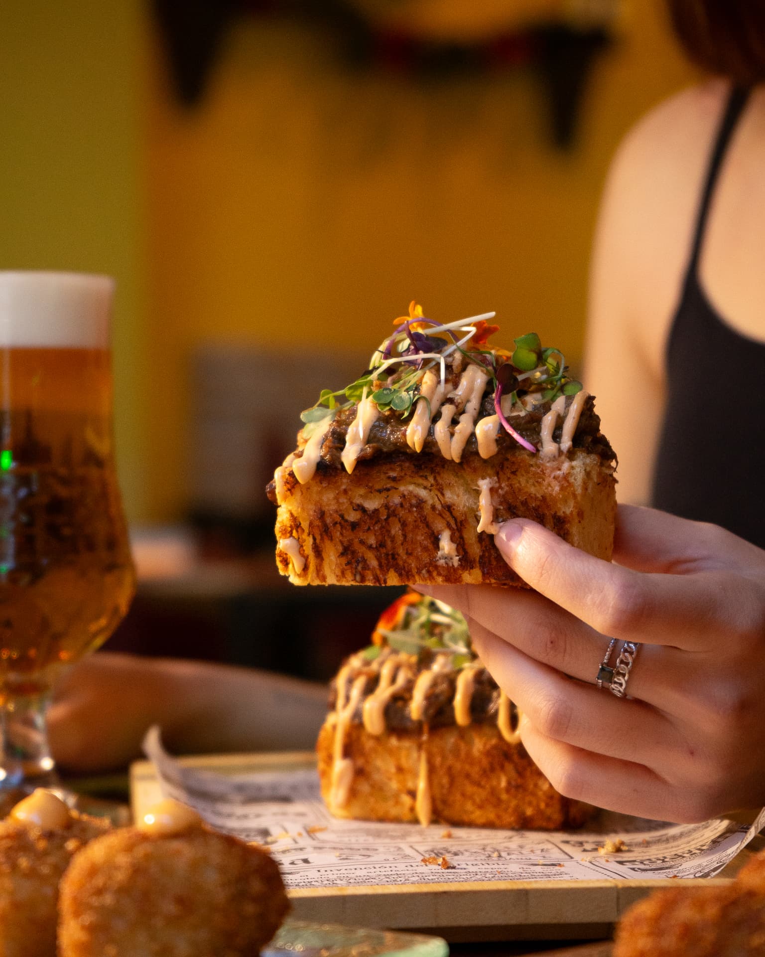 Mano sosteniendo una tosta con carne y brotes junto a una cerveza